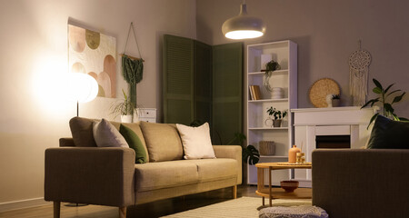 Fototapeta premium Interior of stylish living room with grey sofas, coffee table, fireplace and glowing lamps at evening