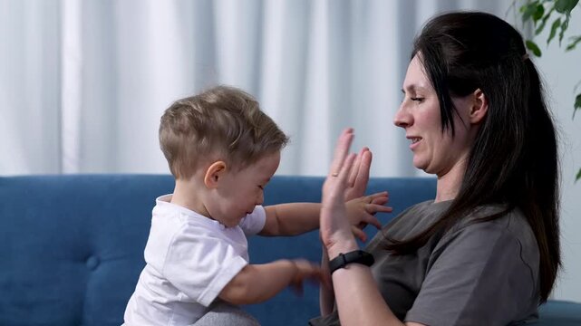 Mother and toddler playing clapping game at home. Mom claps hands with small child on blue sofa. Parent and baby bond together. Happy mother plays with son indoors. Family play on cozy couch at home.