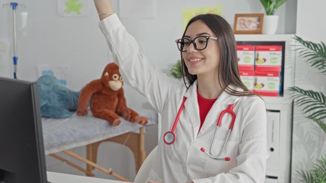 Hispanic woman doctor raising right arm with stethoscope at building desk, smiling at computer; pediatric care warmth.