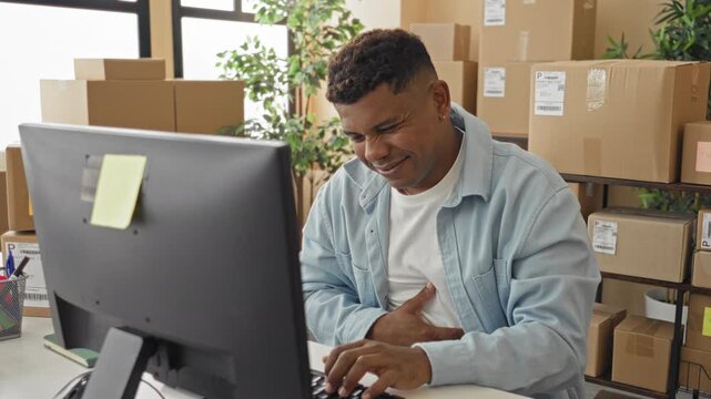 Man clutching chest at computer amid stacked packages in a packing building, grimacing while typing orders, possible heartattack; distress urgency help.