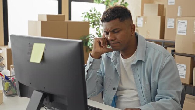 Man using computer with hand on chin and rubbing temples, surrounded by stacked parcel boxes and shipping labels inside a building; frustration.