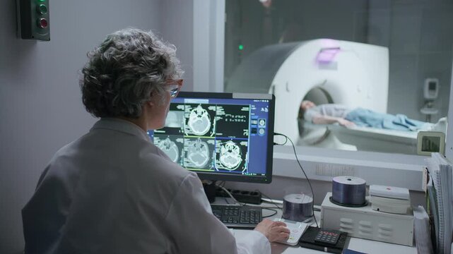 Senior female radiologist sitting in control room, reviewing brain CT scans on computer screen, taking notes and monitoring patient undergoing tomography examination through window
