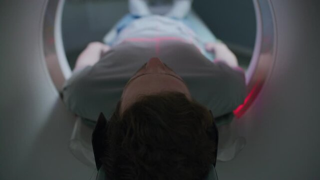Male patient lying on sliding bed, entering CT scanner with red laser lines marking his torso during diagnostic imaging procedure at clinic. View from behind his head