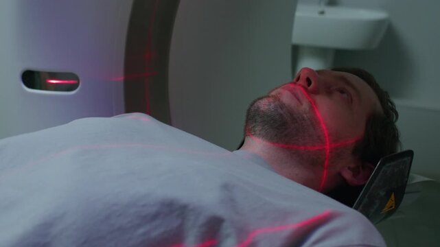 Young male patient entering medical scanner on sliding bed as red laser lines positioning across his face during computed tomography examination in hospital. Close up shot