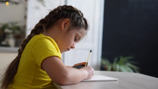 Girl writing in notebook at desk. Child doing homework with pen. Focused girl studying at home. Kid writing notes in spiral notebook. Young student learning. Child concentrating on schoolwork.