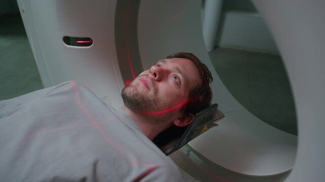 Young male patient lying on sliding bed as entering tomography scanner, red laser lines projected across his face and torso during medical procedure at clinic