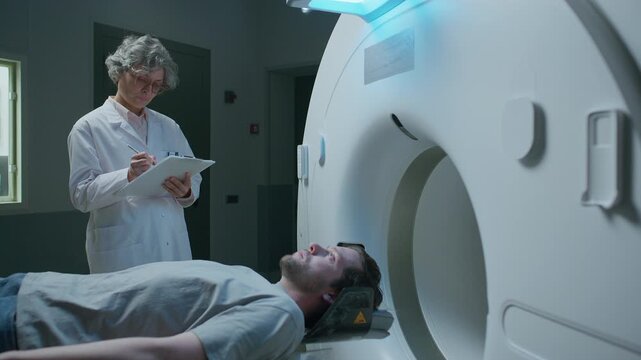 Middle aged female physician writing on clipboard while talking to young male patient lying on sliding bed of advanced CT scanner machine, preparing him for diagnostic procedure at clinic