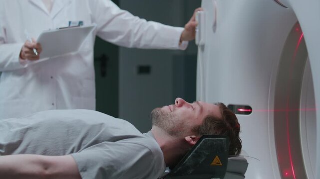 Patient lying on motorized table as sliding out of modern CT scan machine, doctor in white coat standing by with clipboard and operating control panel during diagnostic procedure at medical clinic