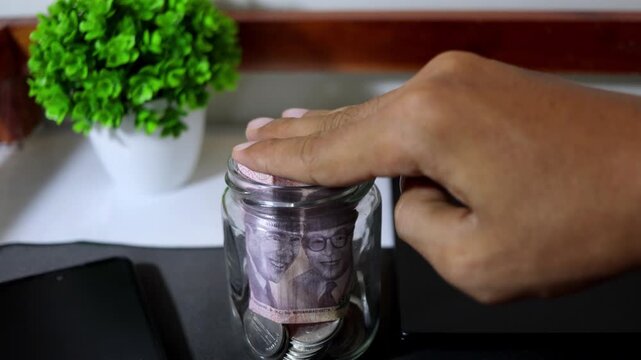 Close up of a man's hand putting an Indonesian Rupiah 100,000 banknote into a glass savings jar on a desk.