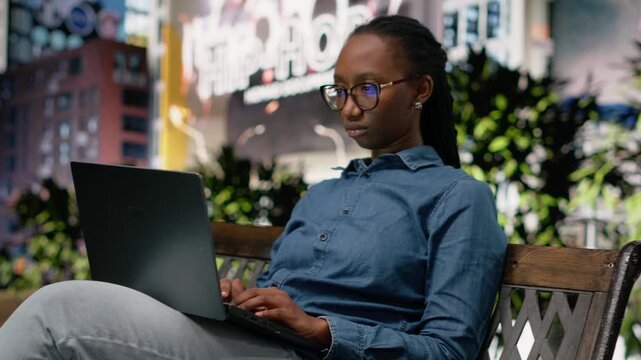 African american woman on park bench holding laptop and working, checking data reports to finish her remote online assignment. Young adult solving tasks outside on notebook.