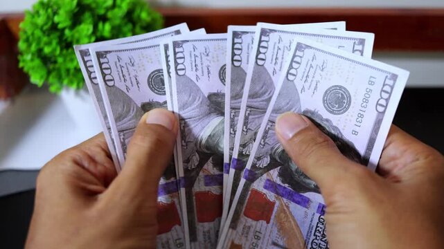 Close up of a man's hands counting one hundred US dollar banknotes on a wooden table.