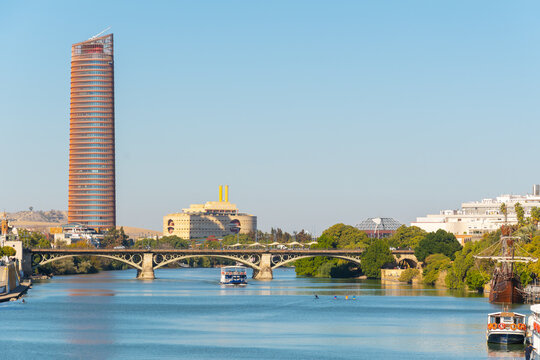 View of the Torre Sevilla skyscraper and the Triana Bridge spanning the Guadalquivir River, with the Nao Victoria ship historic replica in the foreground, in Seville, Spain.
