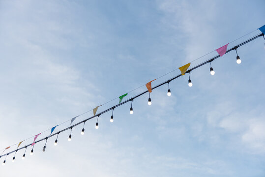 Outdoor party decorations featuring glowing light bulbs and multi-colored triangle flags hanging against  bright blue sky. Perfect for summer festivals, fairs, and outdoor event themes.