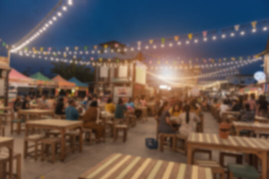 Abstract background of lively night market with people dining at wooden tables under colorful bunting flags and glowing string lights. Ideal for travel and food culture themes.
