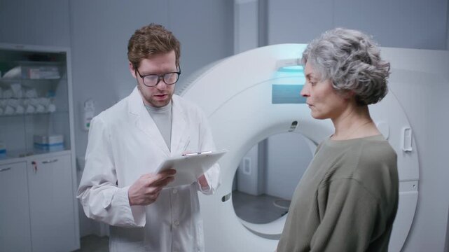 Young male doctor in white lab coat filling out medical history form while talking with senior female patient after CT scan procedure in radiology room