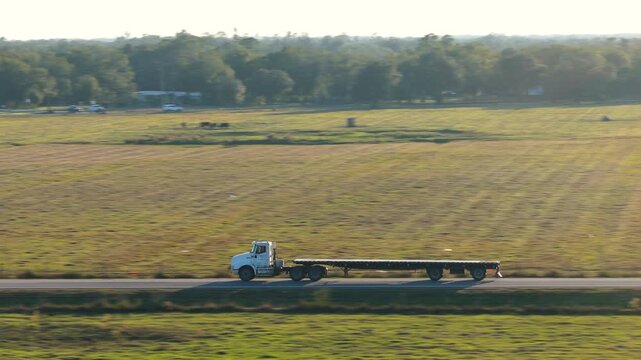 Two lane road through open fields with flatbed truck tractor trailer moving forward at golden hour. Cargo hauling and long distance delivery concept.