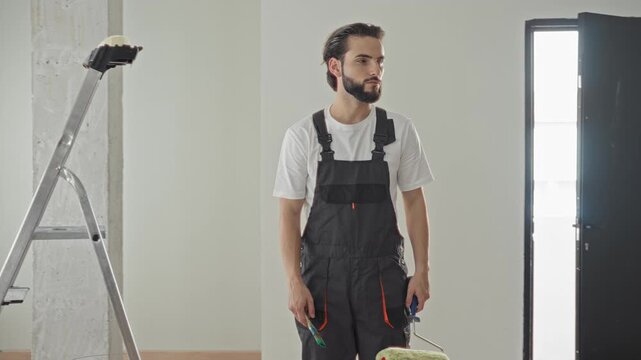 Man painter in overalls holds paint roller and brush looking sideways in building; confidence home renovation.