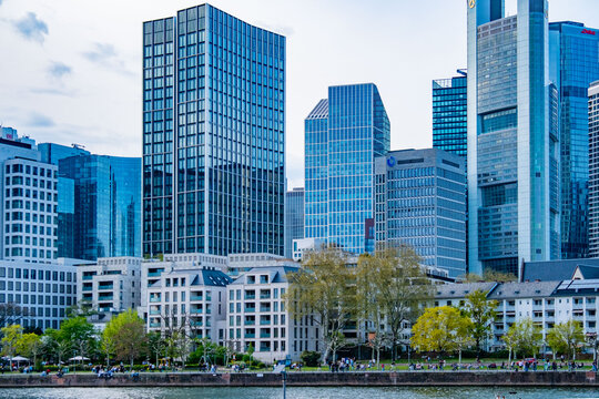 Modern skyline Frankfurt am Main, mainhattan glass skyscrapers, commercial buildings reflecting sky, seen from across river Main with green park and people relaxing, financial capital, urban scenery