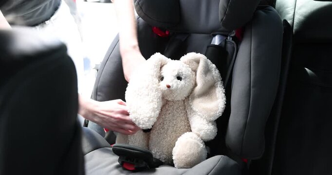 Danger prevention. Woman fastening toy bunny in child safety seat inside car, closeup