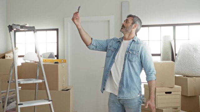 Man in denim shirt holds smartphone up as he surveys packed cardboard boxes, ladder, level and bubble wrap inside a building under renovation; focus organization.