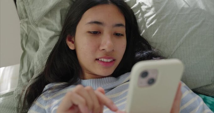 Asian teenage girl lying near on bed, using smartphone, Sunlight filters through sheer curtains in bedroom