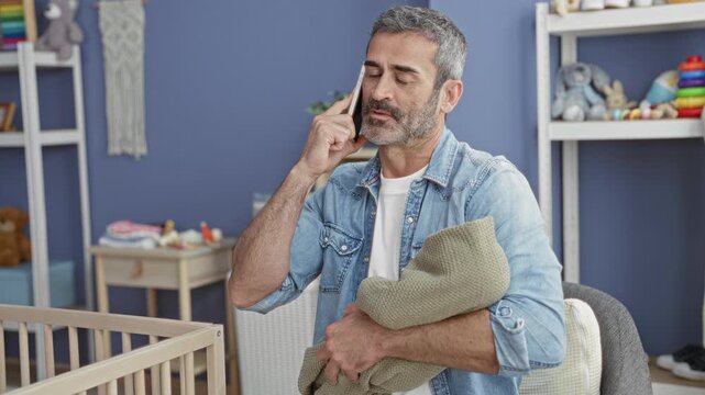 Man holding wrapped baby with visible hand while talking on smartphone beside a wooden cradle and nursery toys in a nursery building; responsibility.