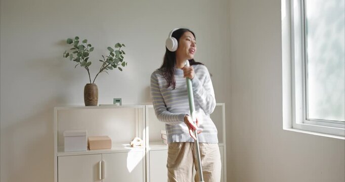 young Asian woman in a striped sweater dances with a mop as a microphone in a bright minimalist home, wearing white headphones