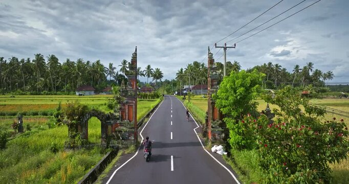 Balinese Candi Bentar split gate standing on a road with a motorcycle and a bicycle passing through, surrounded by lush tropical rice fields and palm trees under a cloudy sky. Travel and explore Bali