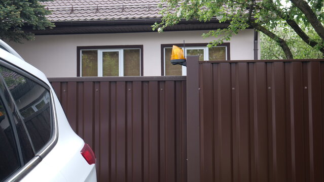 White car exiting a residential property driveway, passing a brown metal fence with an automatic gate warning lamp flashing against the background of a house creating a sense of security and privacy
