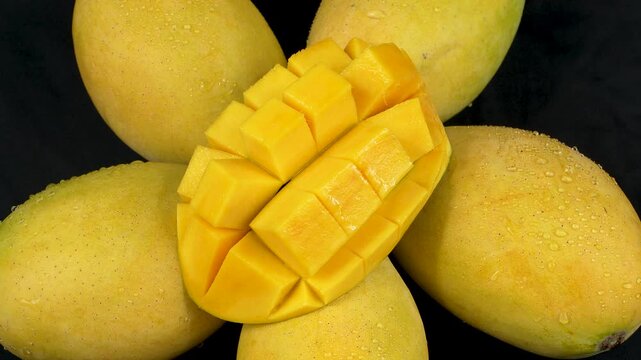 Close-up of a diced yellow mango slice on ripe wet mangoes rotating on a black background.