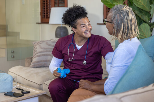 Home RN in scrubs smiling touching Senior African American woman's knee on sofa holding blue gloves