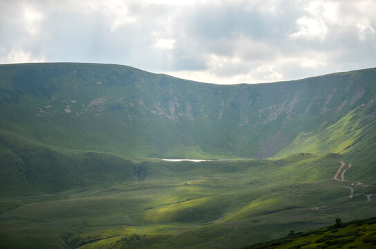 Serene green mountain valley bathed in shafts of sunlight with a small reflective Dogyaska lake and a winding dirt road leading through rolling hills beneath dramatic cloud cover. Carpathians