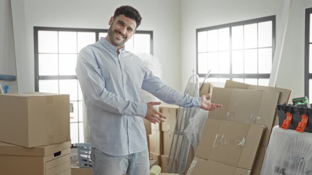 Man, young hispanic, smiling and gesturing with open arms beside stacked cardboard moving boxes and taped parcels in building; contentment fresh start.