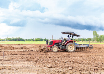 A red agricultural tractor working in a vast rural field, leveling and plowing dry, clumpy soil with a front dozer blade and rear disc harrow under a dramatic, cloudy sky. © Zblaster