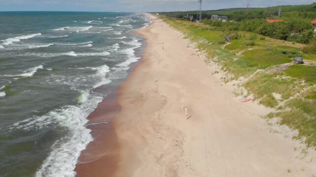 Aerial footage captures the scenic landscape of the Baltic Sea shore near Klaipėda, Lithuania, on a bright summer day. The wide, empty sandy beach meets the gentle waves of the sea, bordered by natura