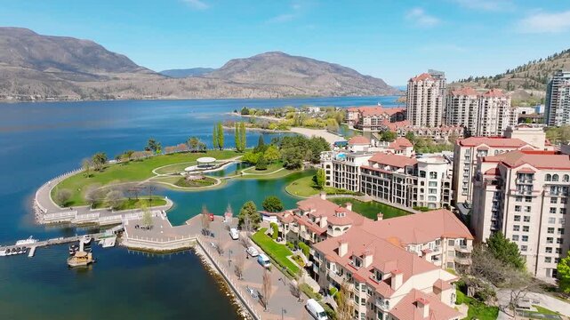 Lakeview of a building complex with coral coloured roofs and man-made landscape at the water's edge, with mountain and water in the background, on a bright and sunny day.