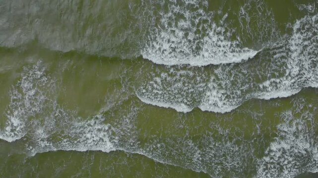 A top down, high angle aerial drone shot showcases the dynamic, textured surface of the Baltic Sea in Lithuania. Powerful emerald green waves crash and churn, creating striking white foam patterns as 
