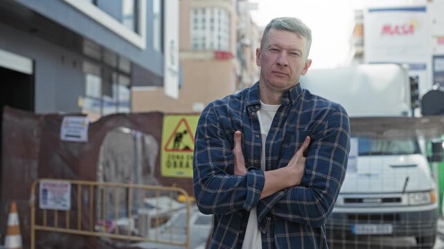 Man arms crossed in front of delivery van at a street construction site, visible hands and plaid shirt; determination.