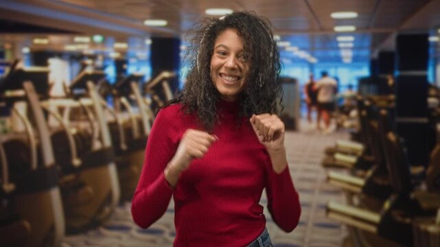 Woman wearing headphones dancing with raised fists and smiling in a fitness studio; joyful upbeat energy.