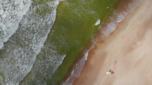 High angle, drone footage captures powerful white capped waves rolling into a vast, sandy coastline. The vibrant green and deep blue ocean water contrasts with the warm, neutral tones of the beach, wh