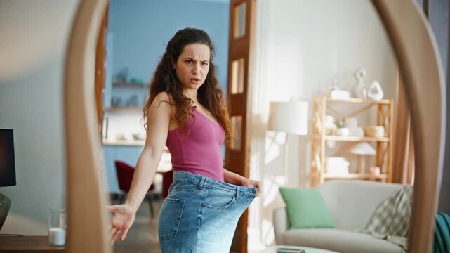 Fit woman examining oversized denim jeans in mirror home. Confused slim brunette
