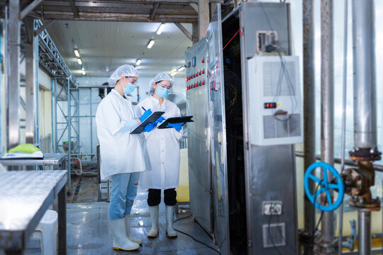 Female quality control team using digital tablet and clipboard for production line audit in modern plant, scientists wearing face masks and safety goggles working in high-standard processing facility