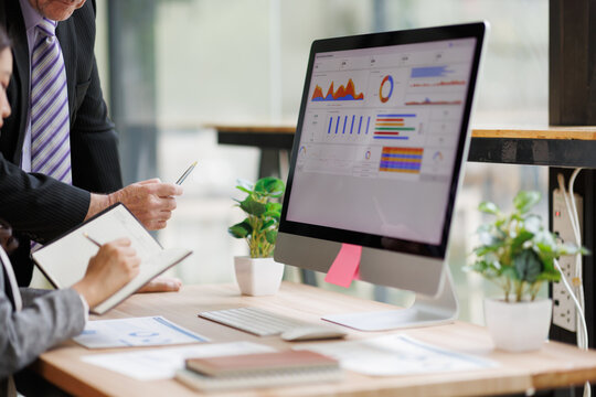 Business professionals analyzing financial data dashboard on a desktop computer, meeting in a modern office, planning strategy and reviewing market reports