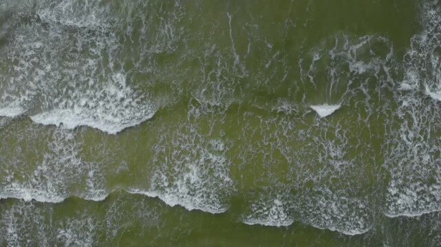 high angle, vertical aerial footage capturing the dynamic movement of emerald green ocean waves as they break and create white foam patterns on the shore of the Baltic Sea in Lithuania.