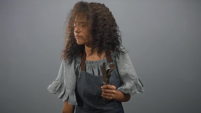 Teenage woman with curly hair wearing denim apron, holding cutlery, finger to lips for silence in studio; secret hush.