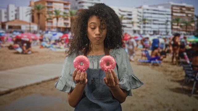 Woman cook holds two pink doughnuts in hands, purses lips and shrugs while facing a seaside building crowded with umbrellas and sunbathers; playful uncertainty.