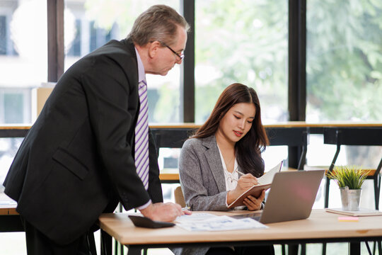 Senior business leader mentoring young female employee at modern office desk, providing guidance, sharing knowledge, and discussing project strategy during working session