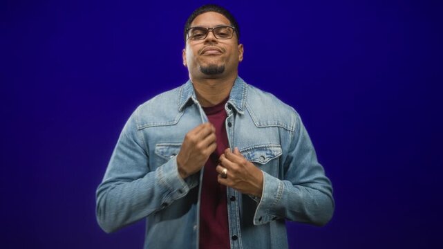 Man with glasses and beard adjusts denim jacket and buttons with both hands in a studio purple set, direct gaze and composed posture; confidence.