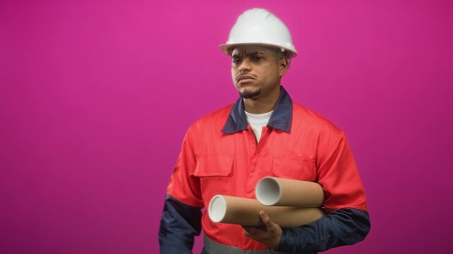 Man construction worker in hard hat holding rolled blueprints and pointing finger in studio; determination focus professionalism integrity.
