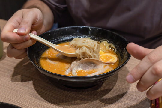 Close-up of a hand holding a spoon with ramen noodles above Tonkotsu broth with chashu pork and ajitama egg in a traditional Japanese ramen bowl at a restaurant.